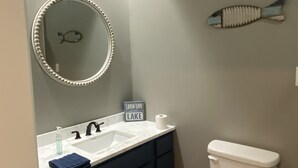 Ein Badezimmer mit einem weißen Waschbecken, dunklen Schränken, einer Toilette, einem Spiegel mit Fischdekor und einem Schild mit der Aufschrift 'Lovin' Life Lake'.
