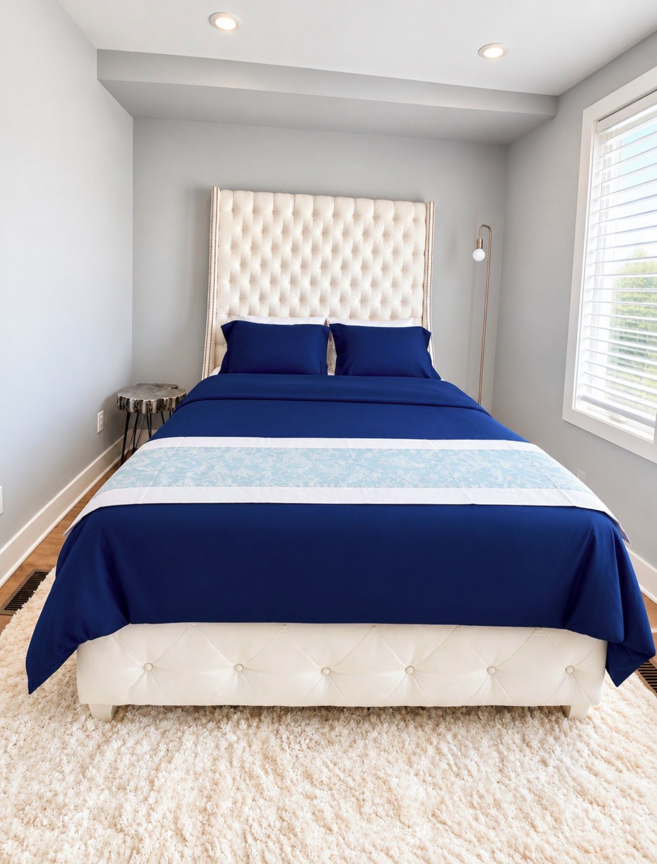 A bedroom with a tufted headboard, a large bed with blue bedding, a small round table, and a floor lamp.