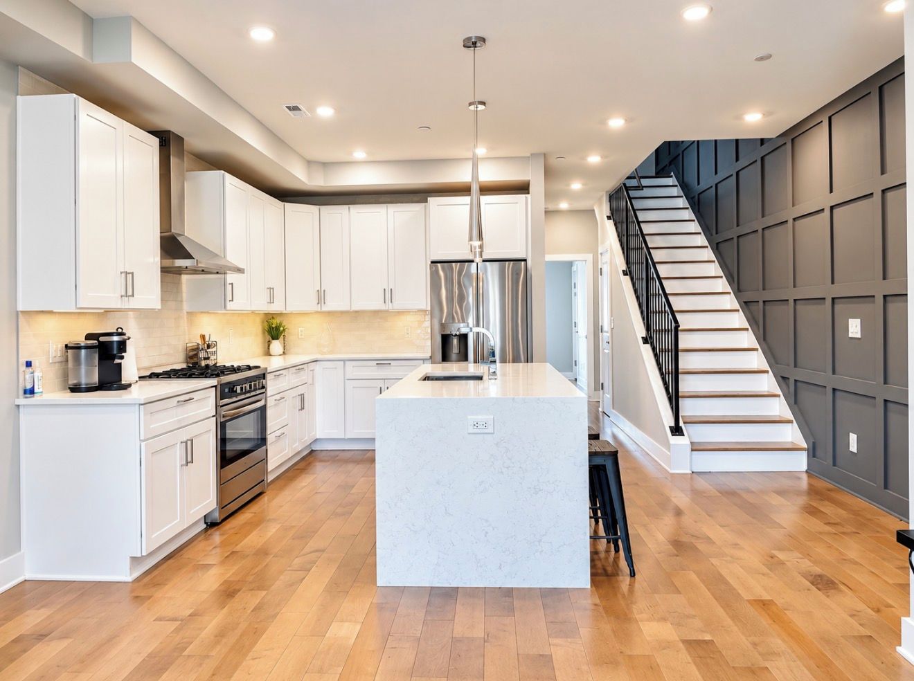 Une cuisine moderne avec des meubles blancs, un îlot central, des appareils électroménagers en acier inoxydable et un escalier en bois.