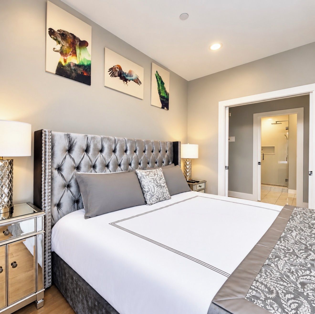 A bedroom with a tufted headboard, a large bed with grey bedding, two bedside tables with lamps, and three wall-mounted artworks.