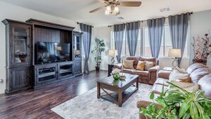 A living room with a dark wood entertainment center, a flat-screen TV, a ceiling fan, and a large window with curtains. - Inviting Sunny House! Poker table~ 4br 3ba 7 beds/ 10 mins to Airport & Strip! (Las Vegas)