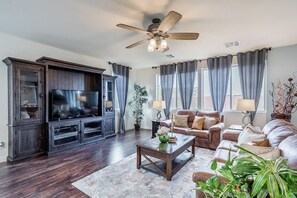 A living room with a dark wood entertainment center, a flat-screen TV, a ceiling fan, and a large window with curtains. - Inviting Sunny House! Poker table~ 4br 3ba 7 beds/ 10 mins to Airport & Strip! (Las Vegas)