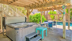A wooden hot tub under a pergola with a pool and patio furniture.