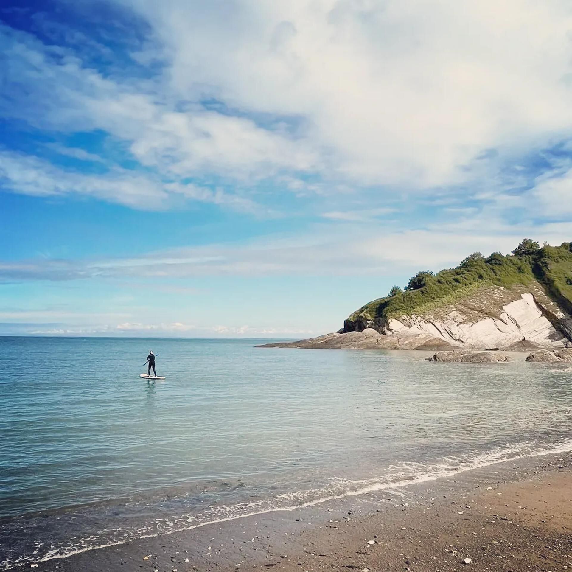 Een strand met helder water, iemand die op een paddleboard staat en een rotsachtige klif met begroeiing.