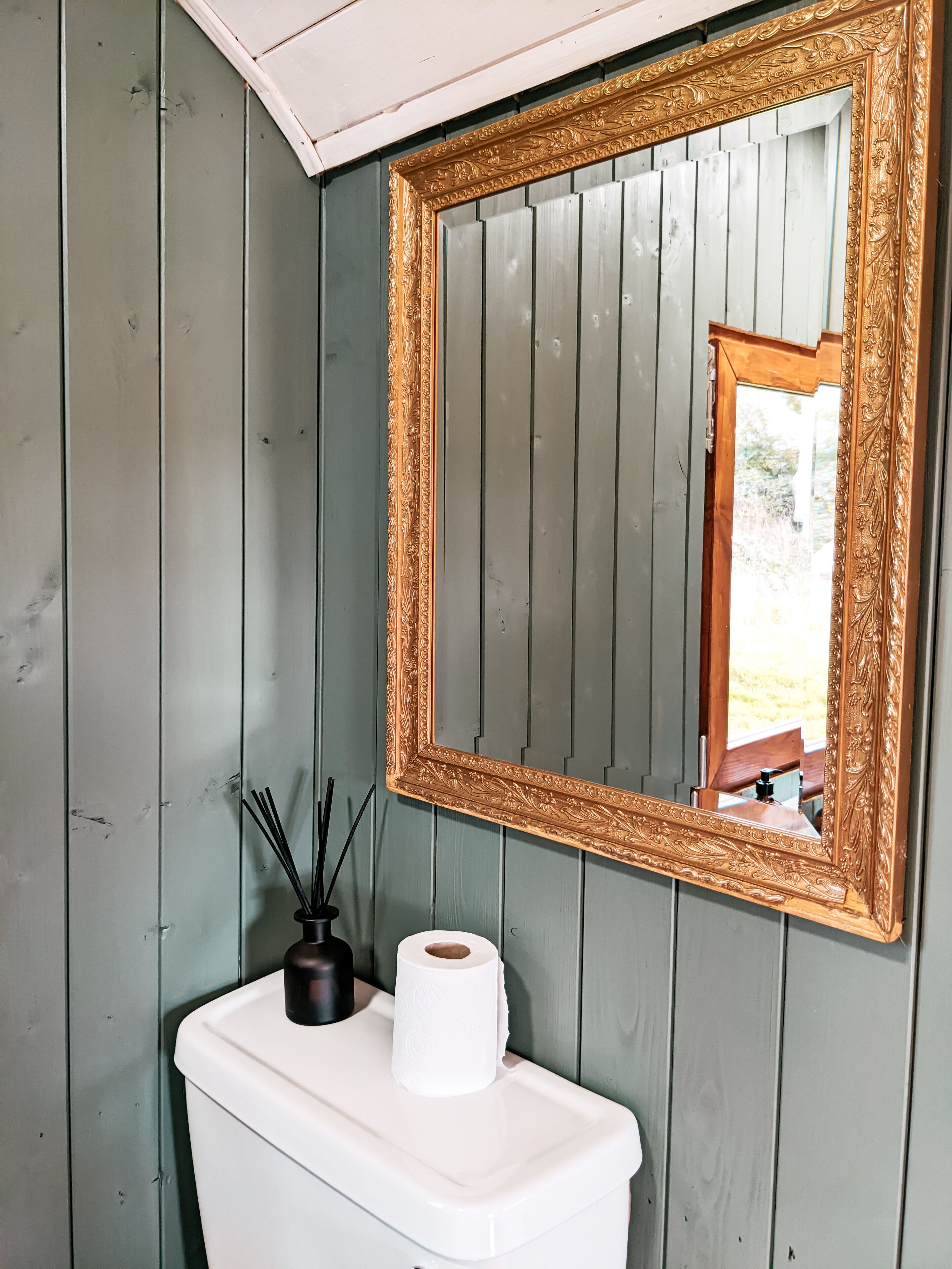 Et badeværelse med toilet, et spejl med udsmykket ramme og en flaske med reededispenser.