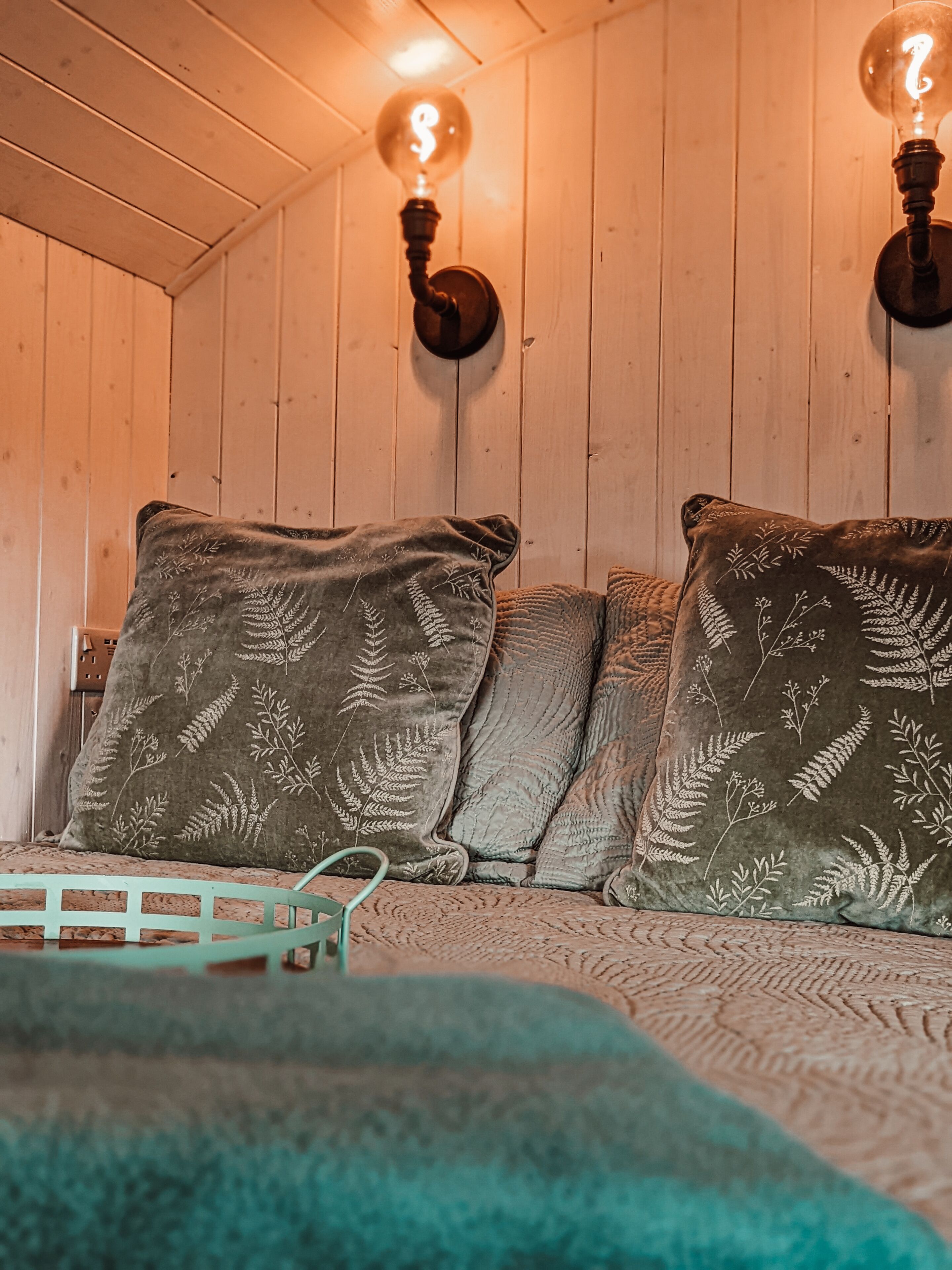 A cozy room with wooden walls, a bed with a green blanket, and two wall-mounted light fixtures with exposed bulbs.