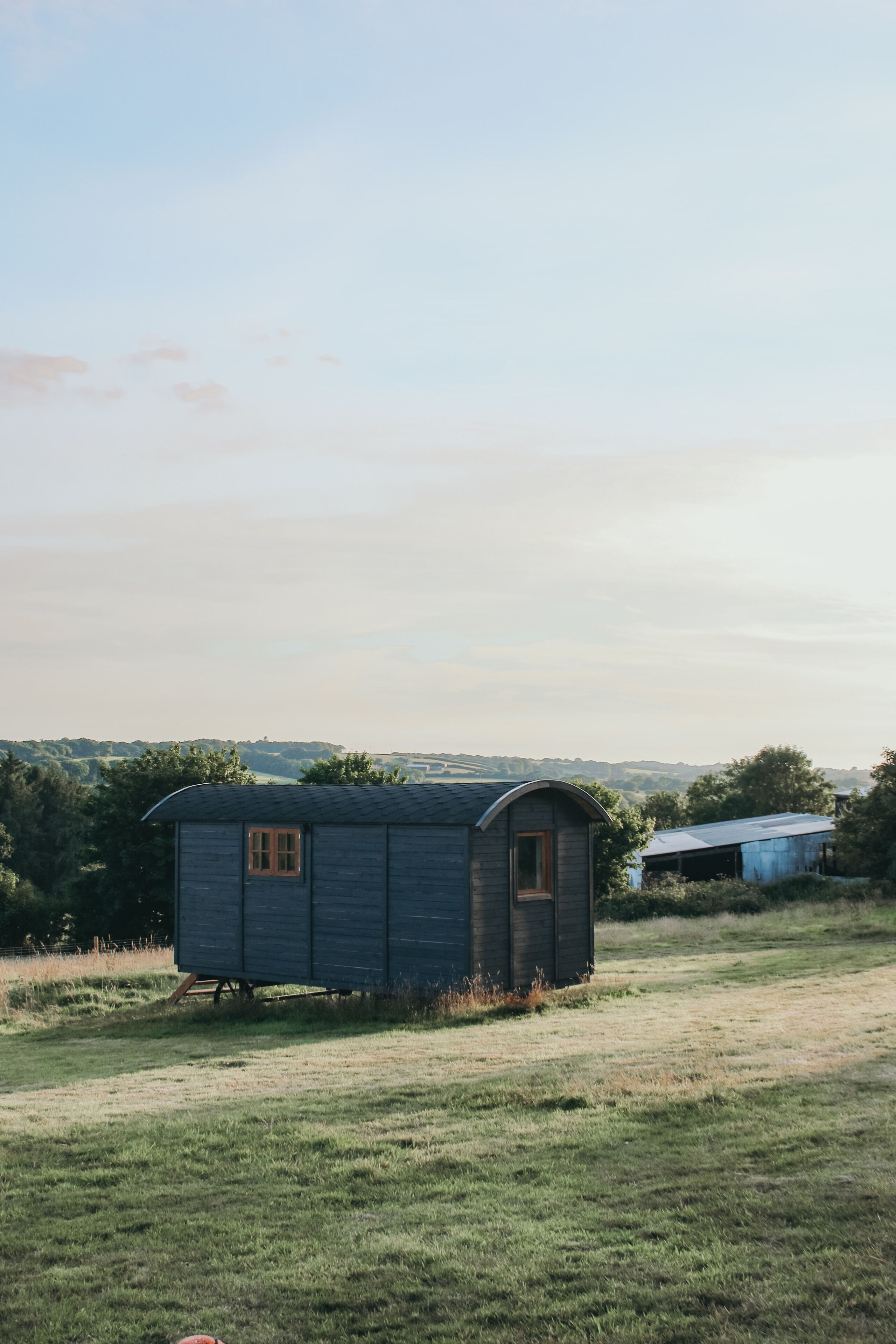 Eine kleine, eingeschossige Holzhütte mit geneigtem Dach, eingebettet in eine Wiese vor Bäumen und Hügeln.