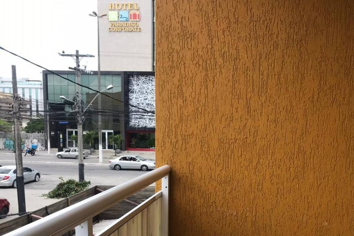 A hotel exterior with a textured wall, a balcony with a metal railing, and a street view with cars and buildings in the background.