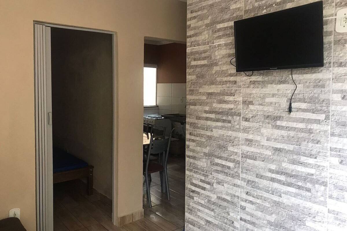 A room with a bed, a TV mounted on a textured wall, and a kitchen area in the background.