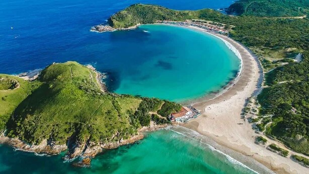 An aerial view of a coastal area with a beach, clear turquoise waters, and green hills. - Jardim Caiçara - Aluguel Economico (Cabo Frio)