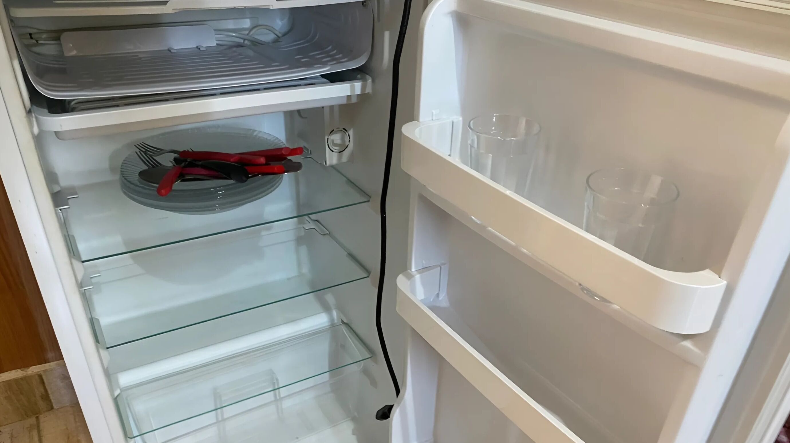 An open refrigerator with clear shelves and a glass door, containing a few items including a red-handled knife and some clear glasses.
