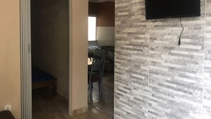 A room with a bed, a TV mounted on the wall, and a kitchen area with a dining table and chairs. - Jardim Caiçara - Aluguel Economico (Cabo Frio)