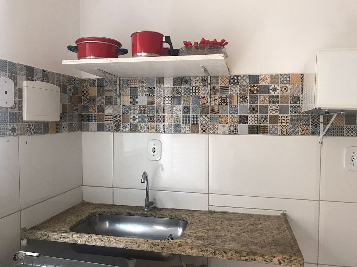 Eine Küche mit einer Granitarbeitsplatte, einer Edelstahlspüle, einer weißen Spritzschutzwand mit gemusterten Fliesen und einem roten Topf auf einem weißen Regal.