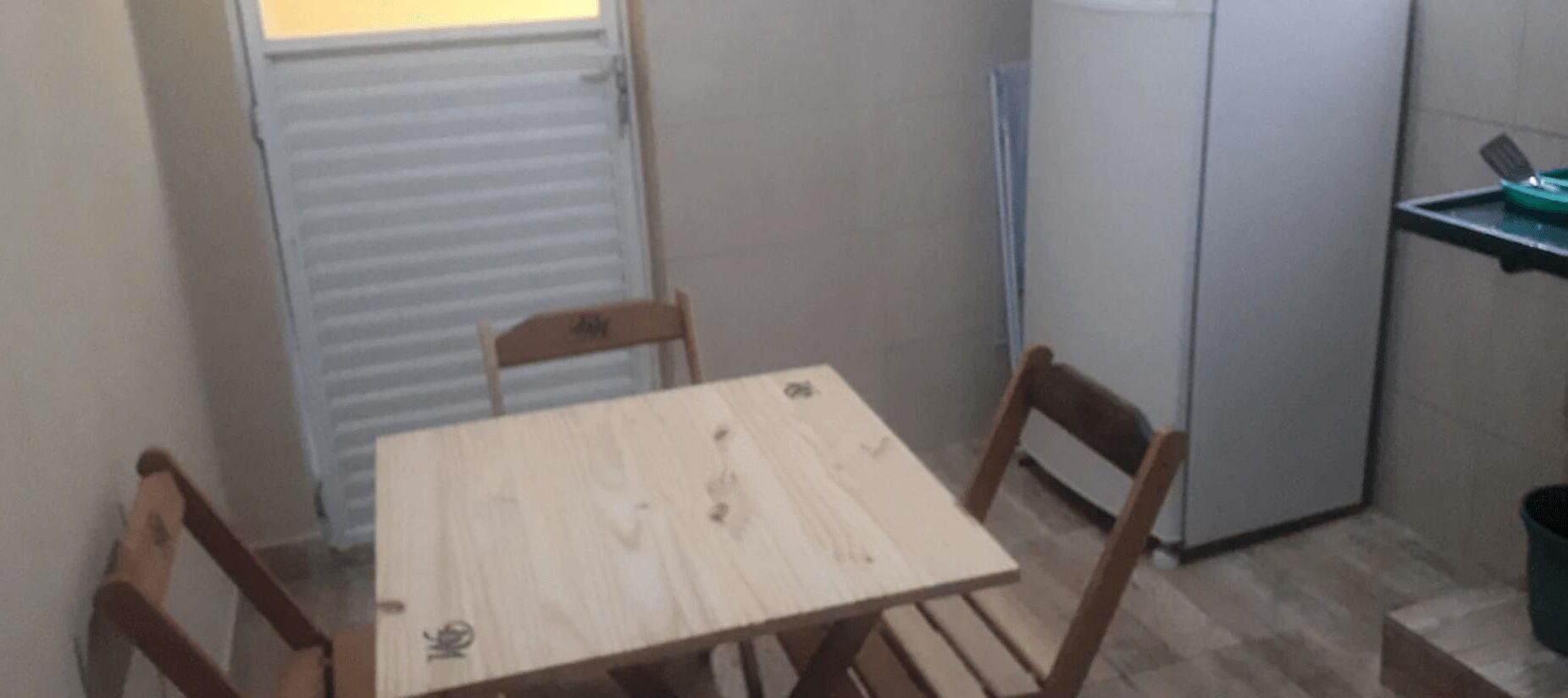 Uma cozinha pequena com uma mesa de madeira e duas cadeiras, uma geladeira e uma janela com persianas.