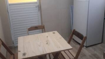Uma cozinha pequena com uma mesa de madeira e duas cadeiras, uma geladeira e uma janela com persianas.