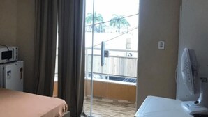 A hotel room with a bed, a microwave, a small refrigerator, a fan, and a view of a balcony with a railing. - Jardim Caiçara - Aluguel Economico (Cabo Frio)