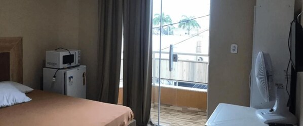 A hotel room with a bed, a microwave, a small refrigerator, a fan, and a view of a balcony with a railing. - Jardim Caiçara - Aluguel Economico (Cabo Frio)