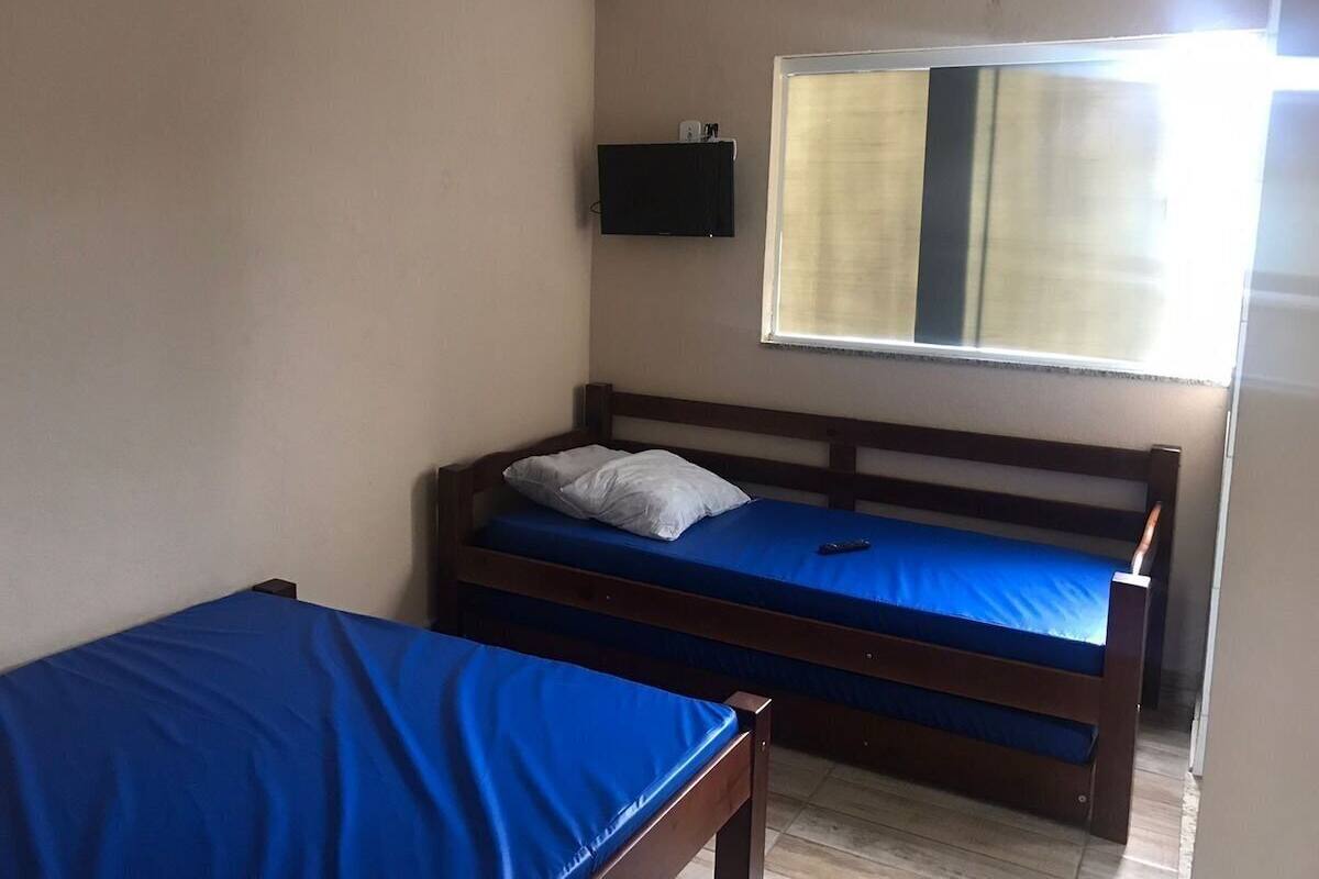 A room with two wooden beds, one with a blue mattress and the other with a white mattress. There is a small black shelf with a television above the bed with the blue mattress.