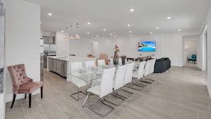 Una cocina moderna con armarios blancos y una isla central, un comedor con mesa de vidrio y sillas blancas, y una sala de estar con sofá y televisor.