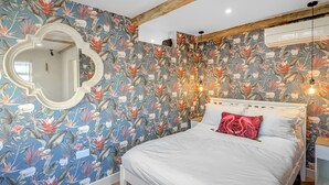 Un dormitorio con papel tapiz floral, una cama blanca, un cojín rojo, techo de madera y aire acondicionado de pared.