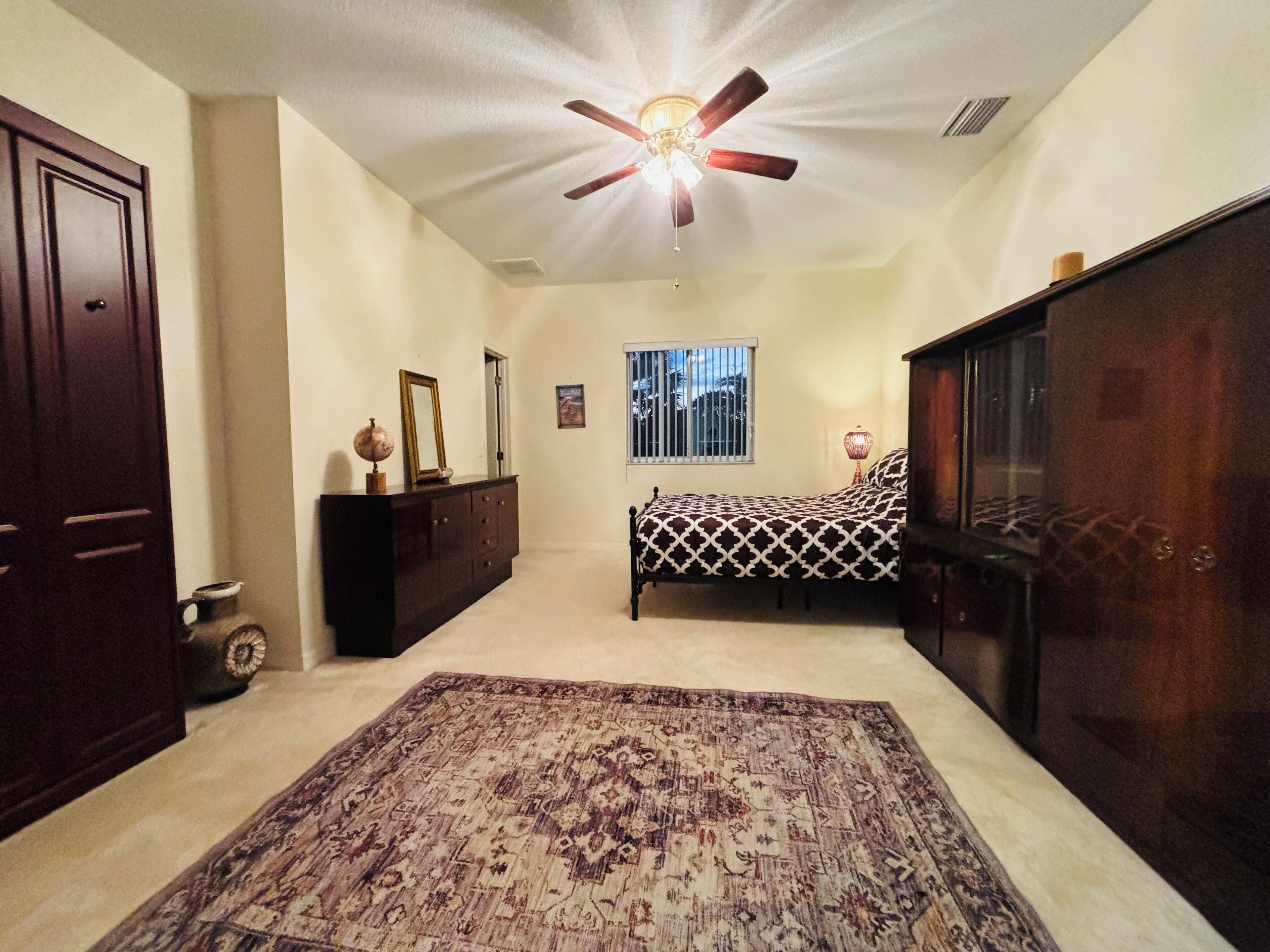 Un dormitorio con cama, cómoda, ventilador de techo, ventana con barrotes y alfombra.