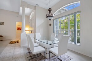 A dining area with a glass table and white chairs, a chandelier, and a large window with a view of palm trees.