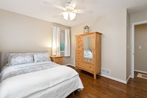 Un dormitorio con cama, cómoda de madera, ventilador de techo y ventana con cortinas.