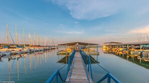 A marina with boats docked, a wooden walkway leading to a covered area, and calm water reflecting the boats.