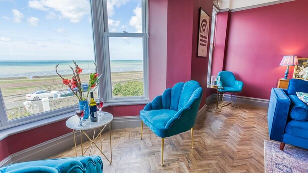 Une pièce dotée d’une grande fenêtre donnant sur une plage, de deux fauteuils bleus, d’une petite table avec des boissons et d’une lampe.