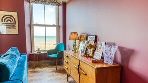 Une pièce avec vue sur la plage, un canapé bleu, une commode en bois ornée d’œuvres d’art et un fauteuil turquoise.
