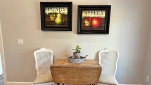 A dining area with two white chairs, a wooden table, and two framed pictures of fruits. - Guest Apartment close to Ski Resorts, Hiking Trails, Shopping Restaurants & more (Holladay)