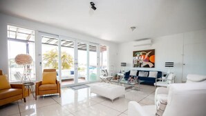 Uma sala de estar moderna com porta de vidro para uma varanda, móveis brancos e um quadro abstrato colorido na parede.