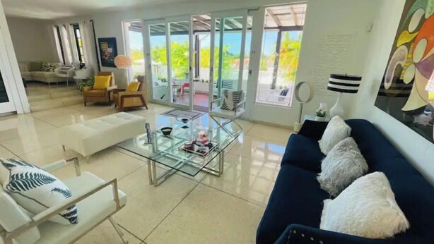 Uma sala de estar moderna com uma mesa de centro de vidro, um sofá azul com almofadas brancas e um sofá branco com almofadas estampadas.