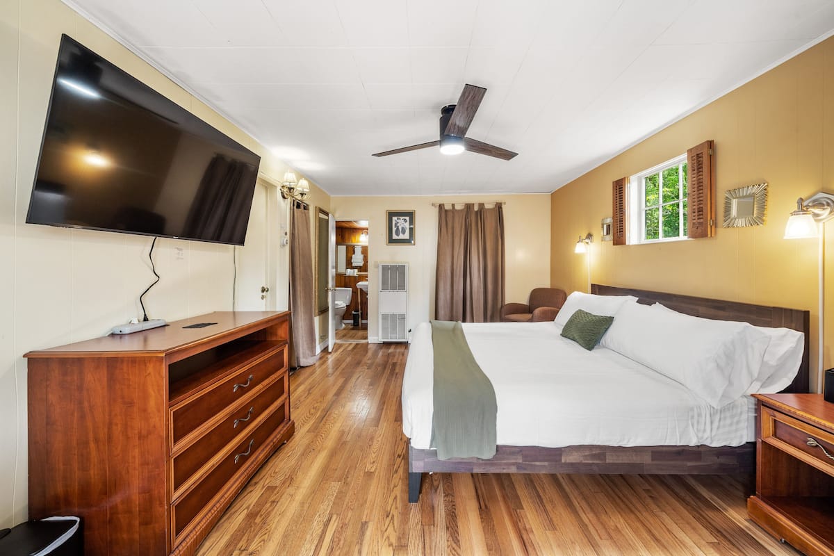 A hotel room with a bed, wooden dresser, TV, ceiling fan, and a bathroom visible in the background.