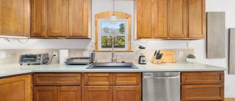 A kitchen with wooden cabinets, a stainless steel dishwasher, and a window with a view of trees.