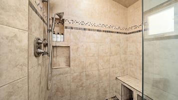 Un baño moderno con baldosas color beige, cabina de ducha de vidrio, banco y estante con artículos de aseo.
