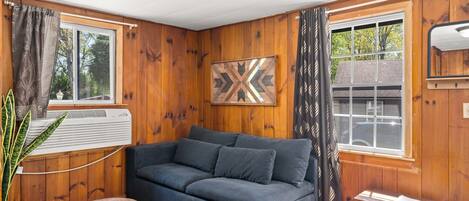 A cozy room with a wooden paneled wall, a grey sofa, a wooden bench, and a window with curtains.