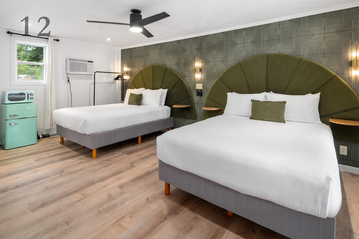 A hotel room with two beds, a ceiling fan, a window with curtains, a wall-mounted air conditioner, and a small green cabinet.