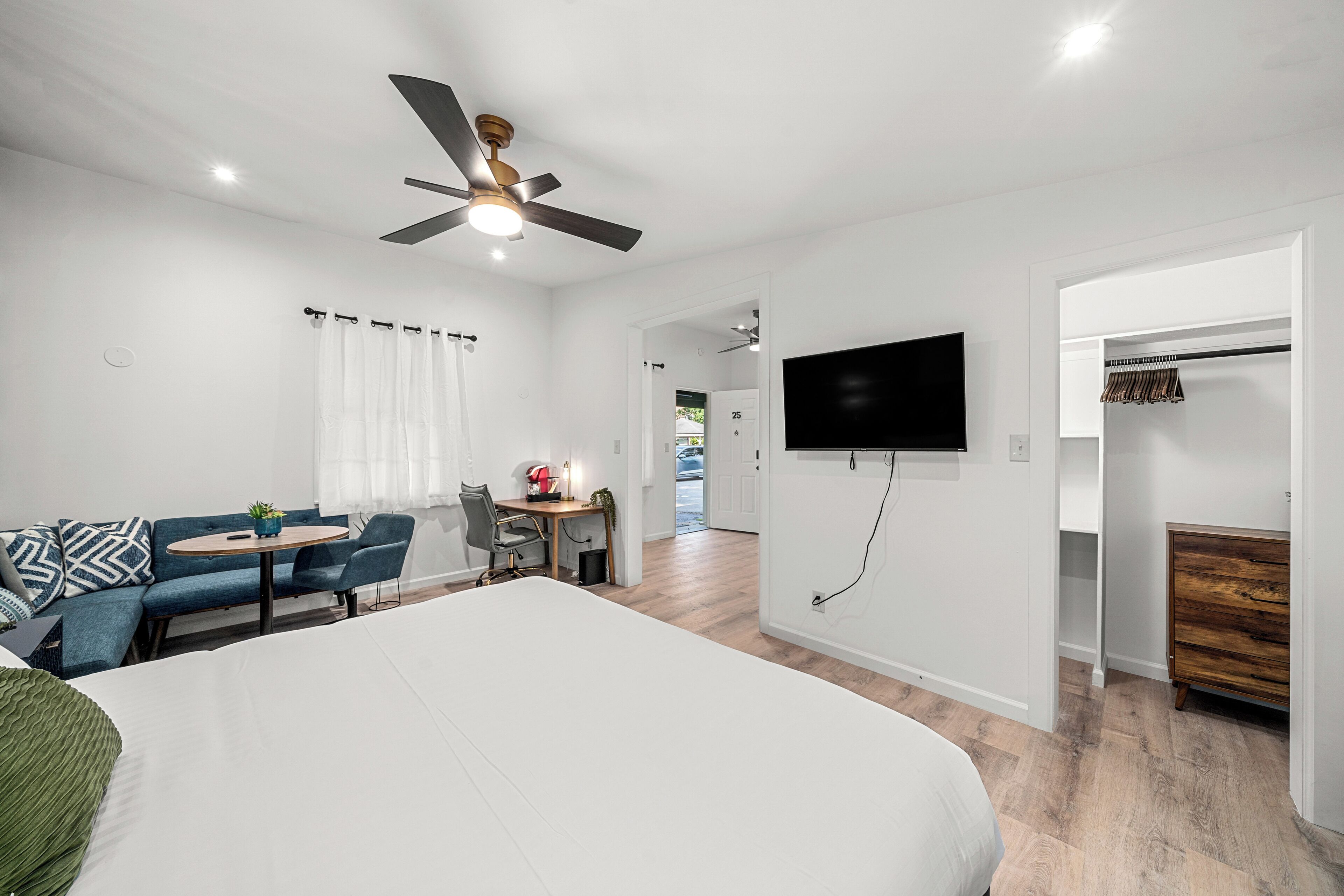 Une chambre moderne avec un grand lit, un ventilateur de plafond, un téléviseur fixé au mur, un bureau avec une chaise et une armoire contenant une commode en bois.