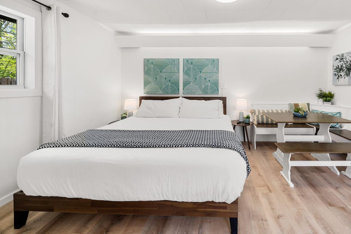 A modern bedroom with a wooden bed, white bedding, a dining table with benches, and two wall-mounted paintings.