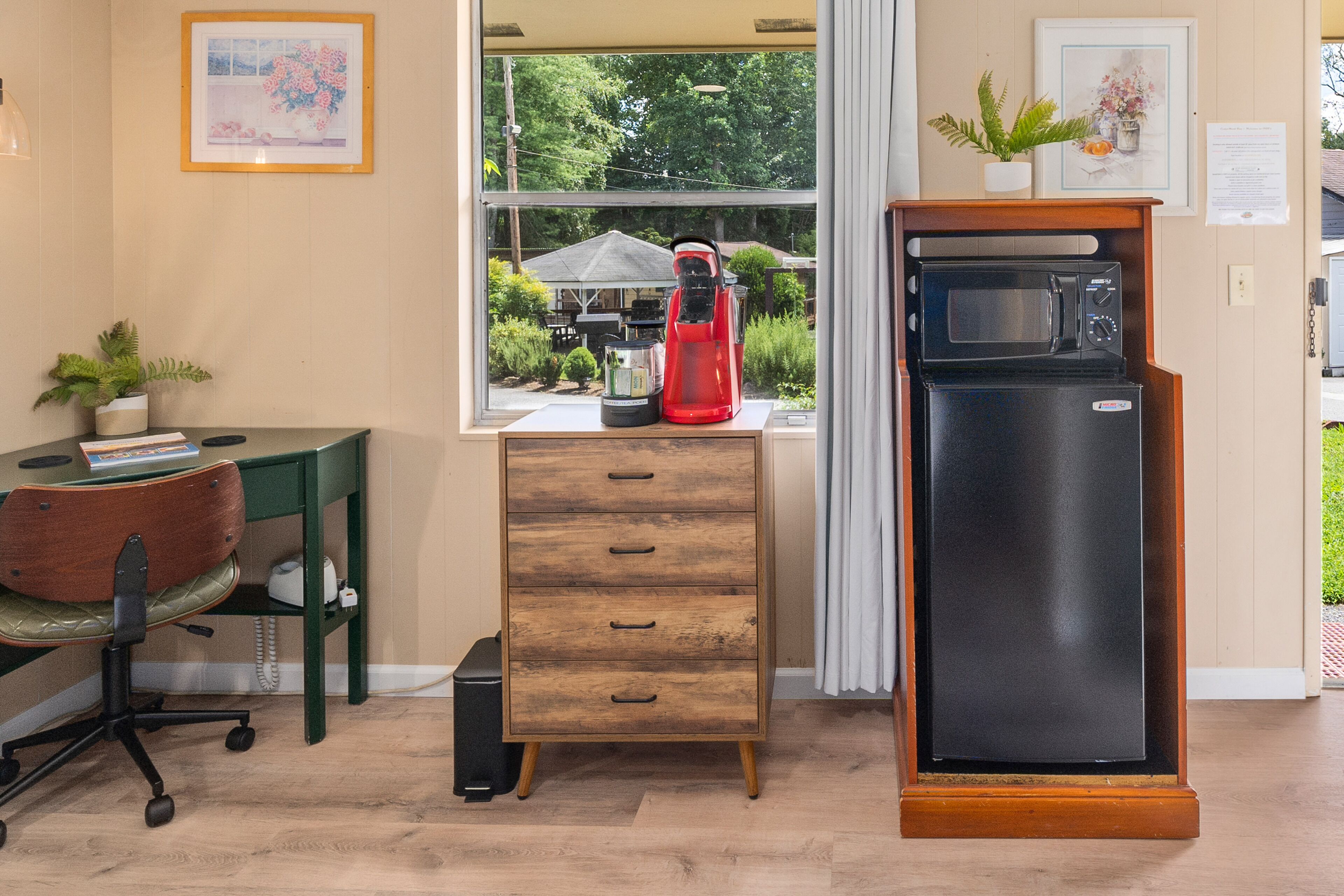 A room with a wooden desk, a red appliance, a black refrigerator, and a window with a view of greenery.