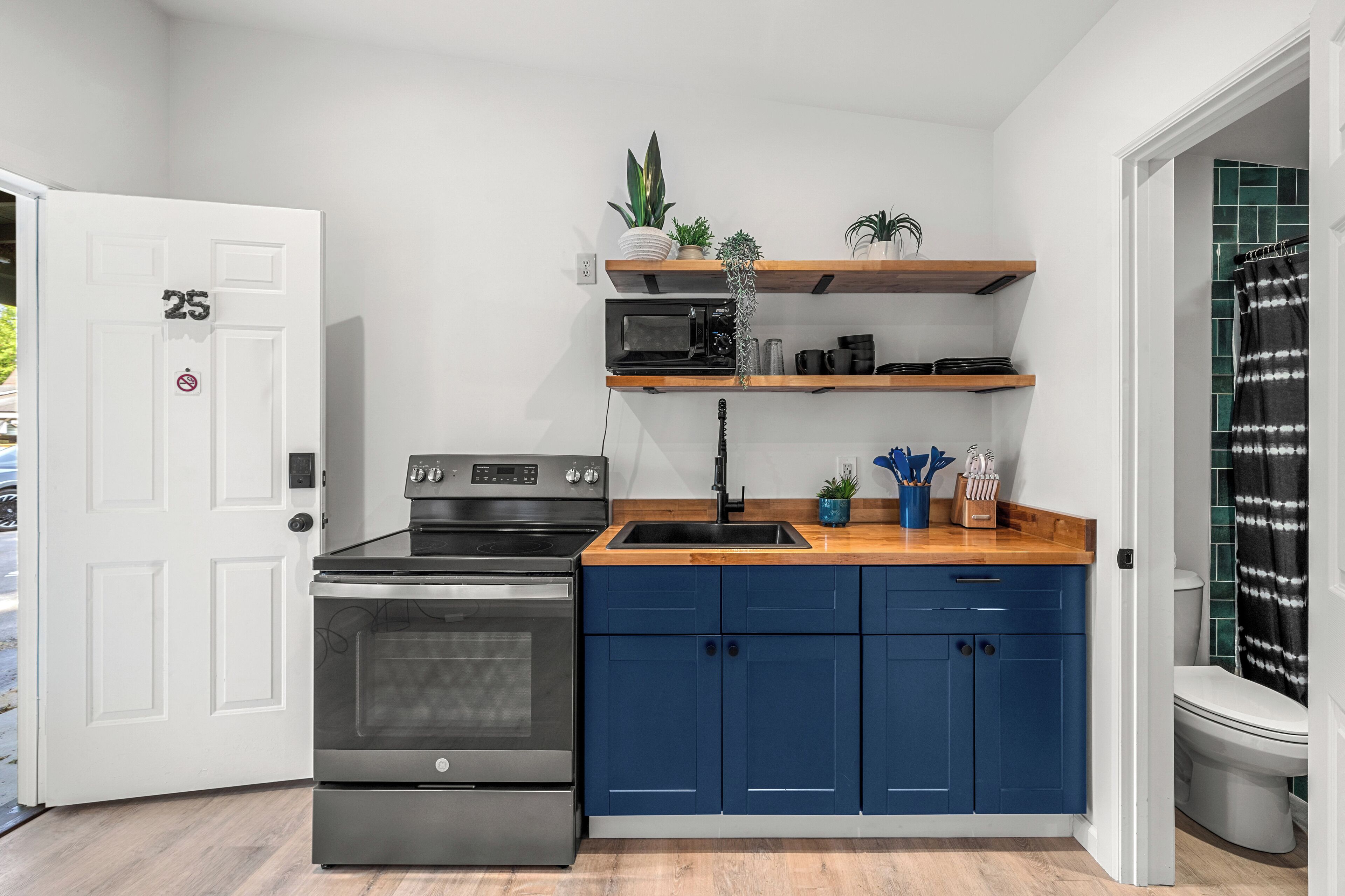 A compact kitchen with a built-in oven, a microwave, and a sink. There are blue cabinets and wooden shelves above the counter.