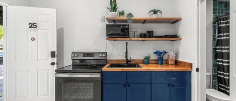 A compact kitchen with a built-in oven, a microwave, and a sink. There are blue cabinets and wooden shelves above the counter.