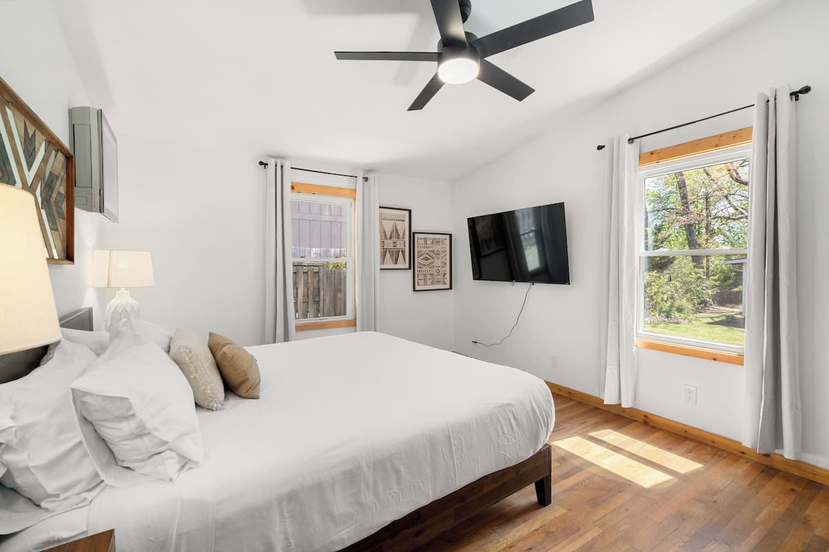 A bedroom with a bed, a ceiling fan, a TV, and large windows with curtains.