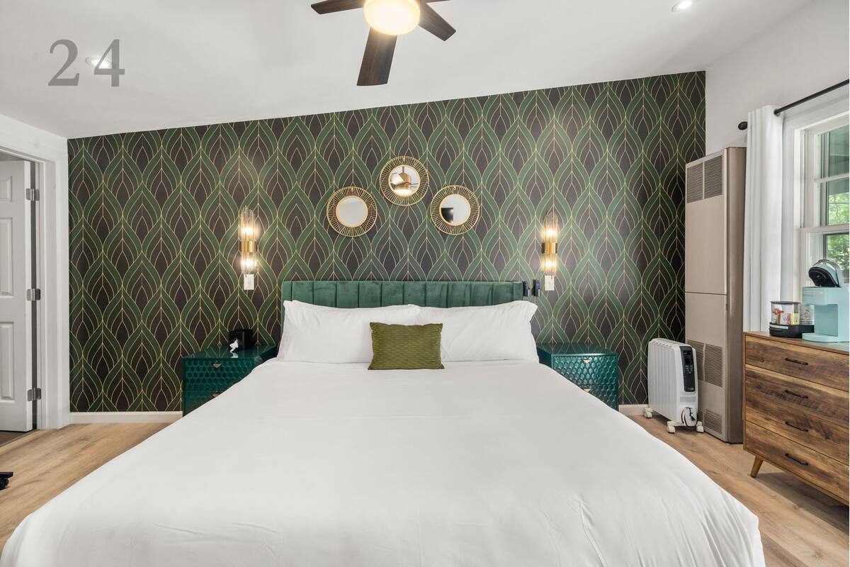 A modern hotel room with a large bed, a green patterned wall, three wall-mounted mirrors, and a ceiling fan.