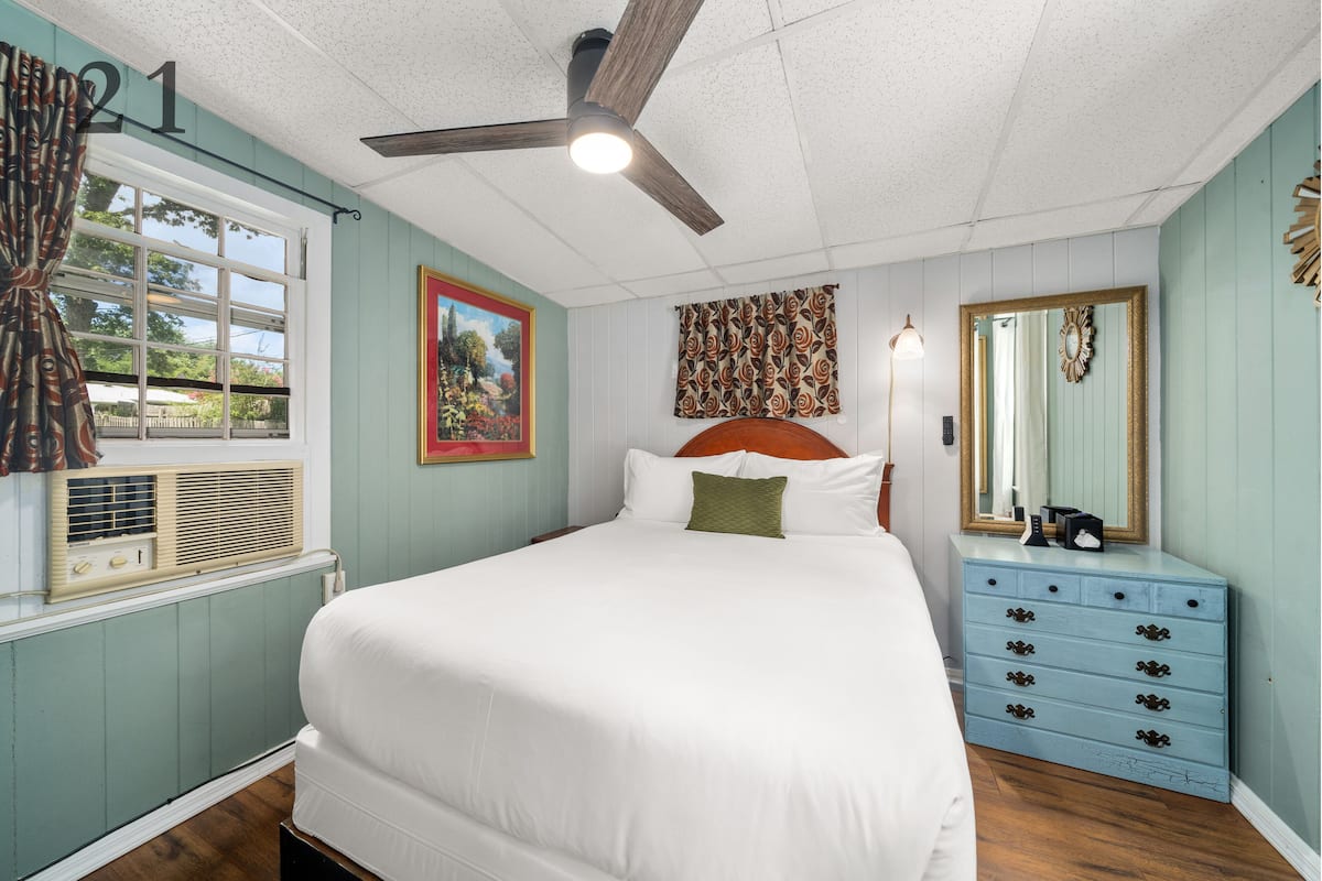 A bedroom with a large bed, a ceiling fan, a dresser, and a window with curtains.