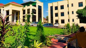 A multi-story building with a flat roof and a covered entrance. There are several cars parked in front, and a person is visible in the foreground. - Golden Castle Hotel (Hoima)