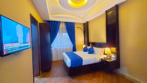 A hotel room with a bed, a TV mounted on the wall, a nightstand with a lamp, and a window with blue curtains. - Golden Castle Hotel (Hoima)