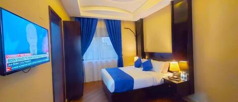 A hotel room with a bed, a TV mounted on the wall, a nightstand with a lamp, and a window with blue curtains.