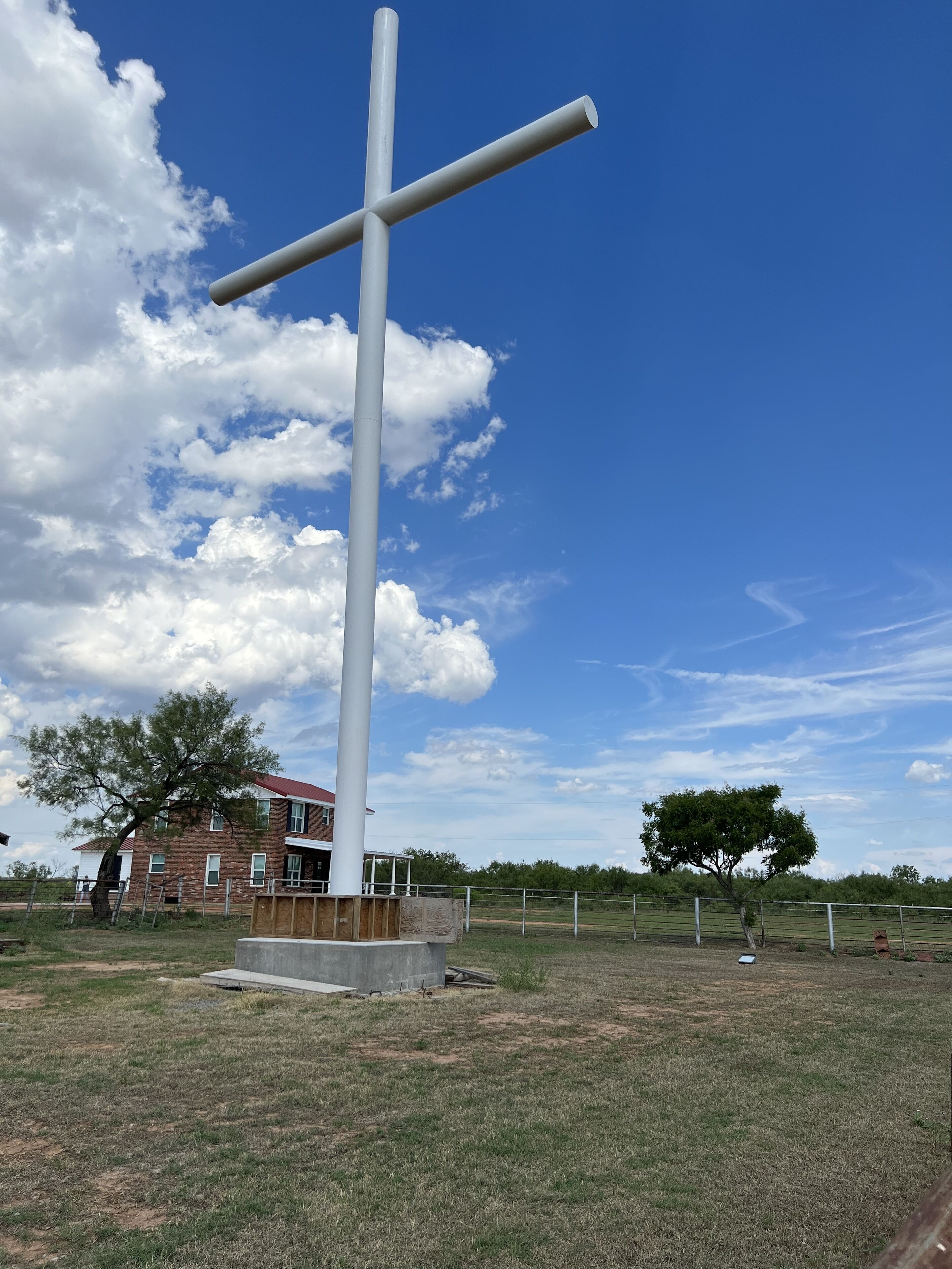 Un alto monumento con forma de cruz blanca en un campo cubierto de césped, con un edificio de ladrillo al fondo.